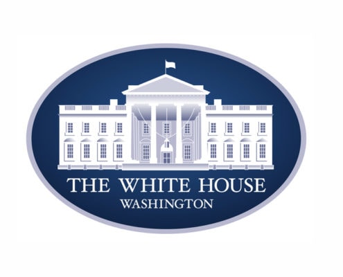 The image shows the official logo of the White House, often seen at events like the Made in America Roundtable. It features a white illustration of the White House on a dark blue oval background, with "THE WHITE HOUSE WASHINGTON" below in capitals.