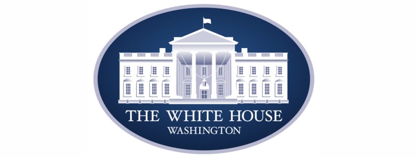The image shows the official logo of the White House, often seen at events like the Made in America Roundtable. It features a white illustration of the White House on a dark blue oval background, with "THE WHITE HOUSE WASHINGTON" below in capitals.