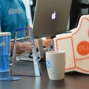 A desk displays a marketing award, a Sideqik tumbler, two logo stickers, a TAG Social Savvy award, a white Sideqik mug, a large foam thumbs-up with "#tagsocialsavvy" and "PGi", and a laptop. In the background, someone wears a blue Sideqik shirt.