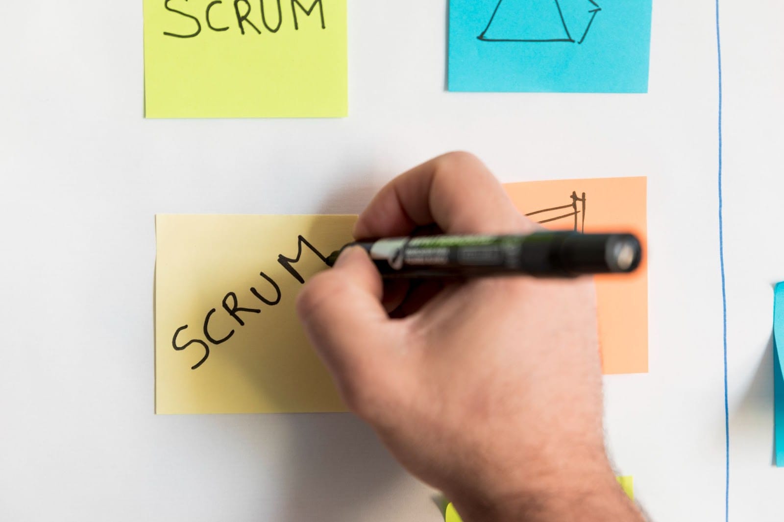 A hand writes the word "SCRUM" in black marker on a yellow sticky note attached to a whiteboard, hinting at Agile software development. Other colorful sticky notes with writing and diagrams suggest an active planning or brainstorming session.
