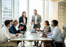 A diverse group of business professionals sits around a conference table in a modern office, discussing and smiling as ABM strategies are shared. Two people stand, leading the Account Based Marketing explained session, while others engage with laptops and take notes.