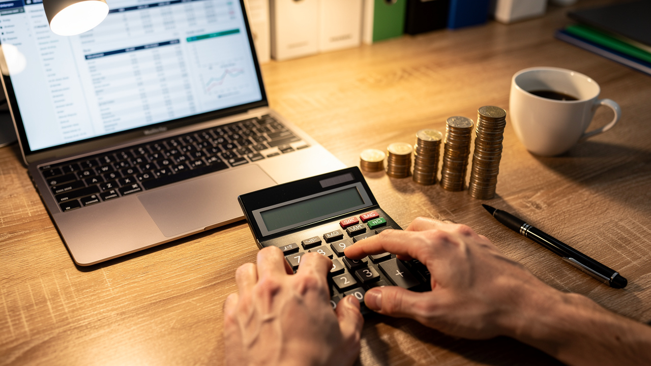 A person uses a calculator at a desk with a laptop displaying financial data, a pen, coffee, and neatly stacked coins, suggesting work related to finance or accounting to measure demand gen and prove impact on demand gen ROI.